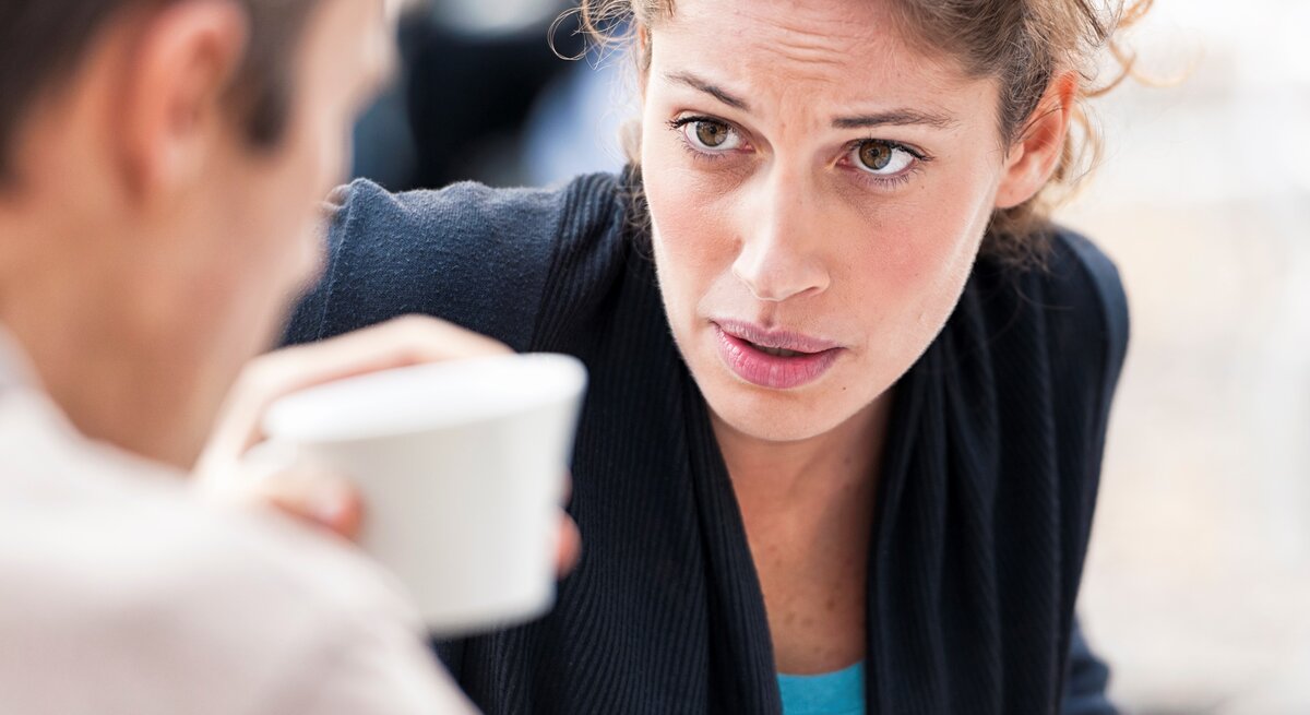Frau schaut im Gespräch im Freien an einem Tisch im ungläubig auf Mann, er hält eine Kaffeetasse