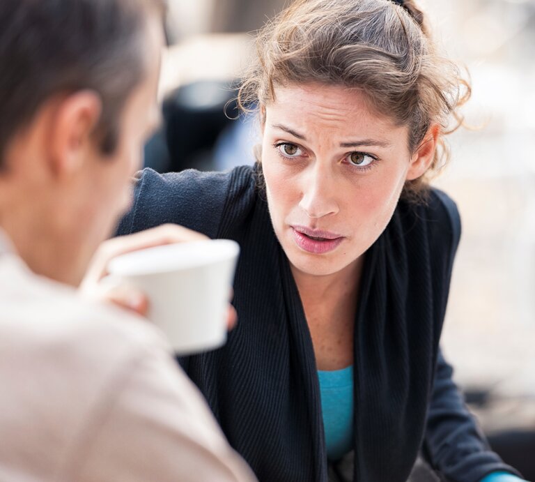 Frau schaut im Gespräch im Freien an einem Tisch im ungläubig auf Mann, er hält eine Kaffeetasse | © Getty Images /	georgeclerk
