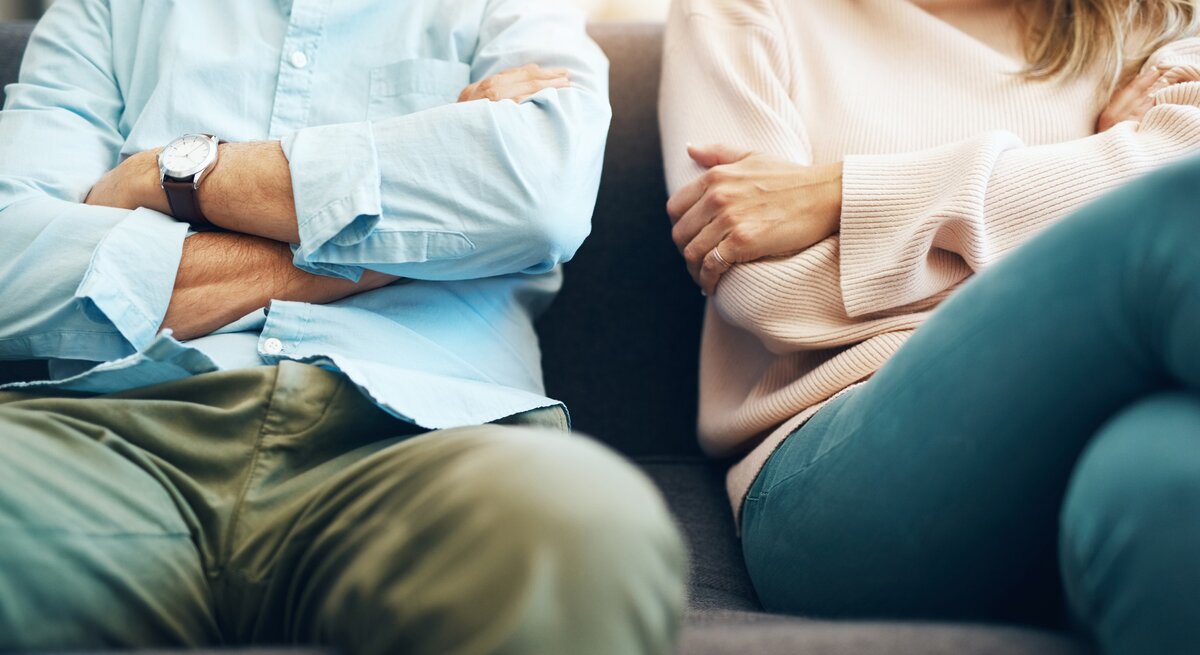 Mann und Frau sitzen nebeneinander mit verschränkten Armen auf einer Couch. Man sieht hauptsächlich den Körper, die Köpfe sind abgeschnitten.