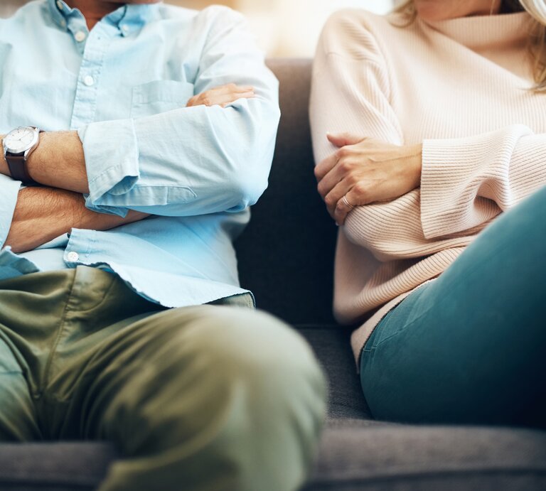 Mann und Frau sitzen nebeneinander mit verschränkten Armen auf einer Couch. Man sieht hauptsächlich den Körper, die Köpfe sind abgeschnitten. | © iStock/PeopleImages