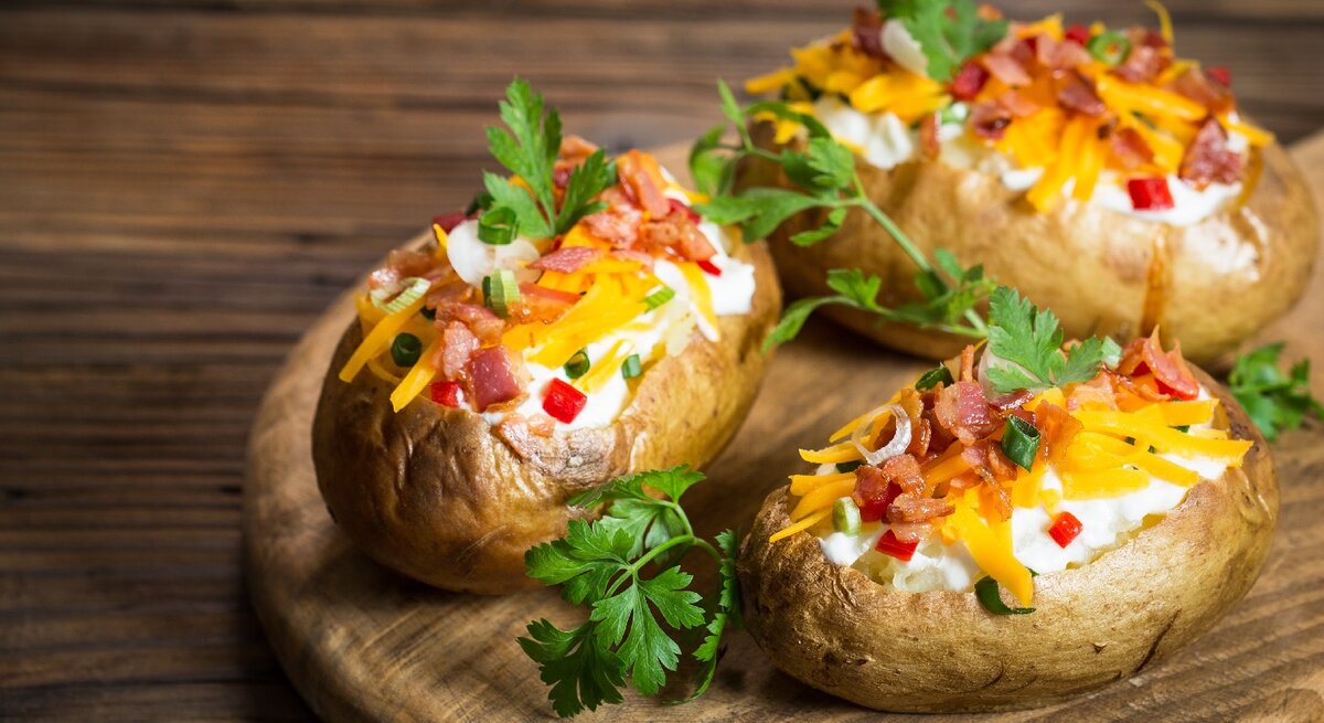 Drei Ofenkartoffeln auf einem Holzbrett. Die Kartoffeln sind gefüllt mit Kräuterquark und mit Petersilie, Käse und Speck bestreut.
