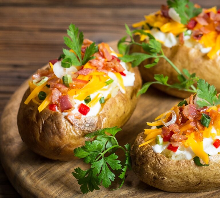 Drei Ofenkartoffeln auf einem Holzbrett. Die Kartoffeln sind gefüllt mit Kräuterquark und mit Petersilie, Käse und Speck bestreut. | © Shutterstock/ pilipphoto