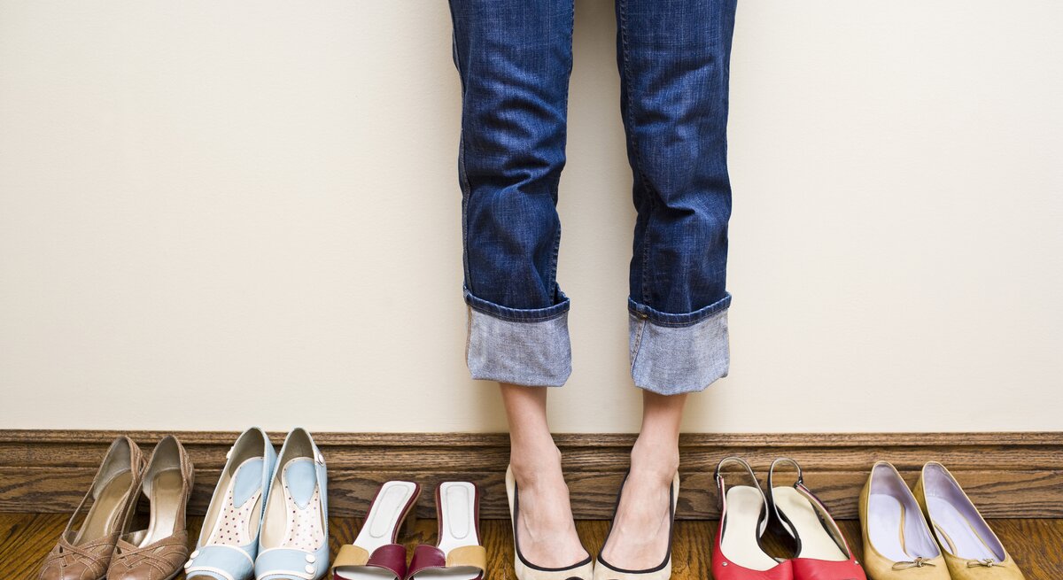 Viele Schuhe in einer Reihe aufgestellt, dazwischen die Beine und Füße einer Frau. 