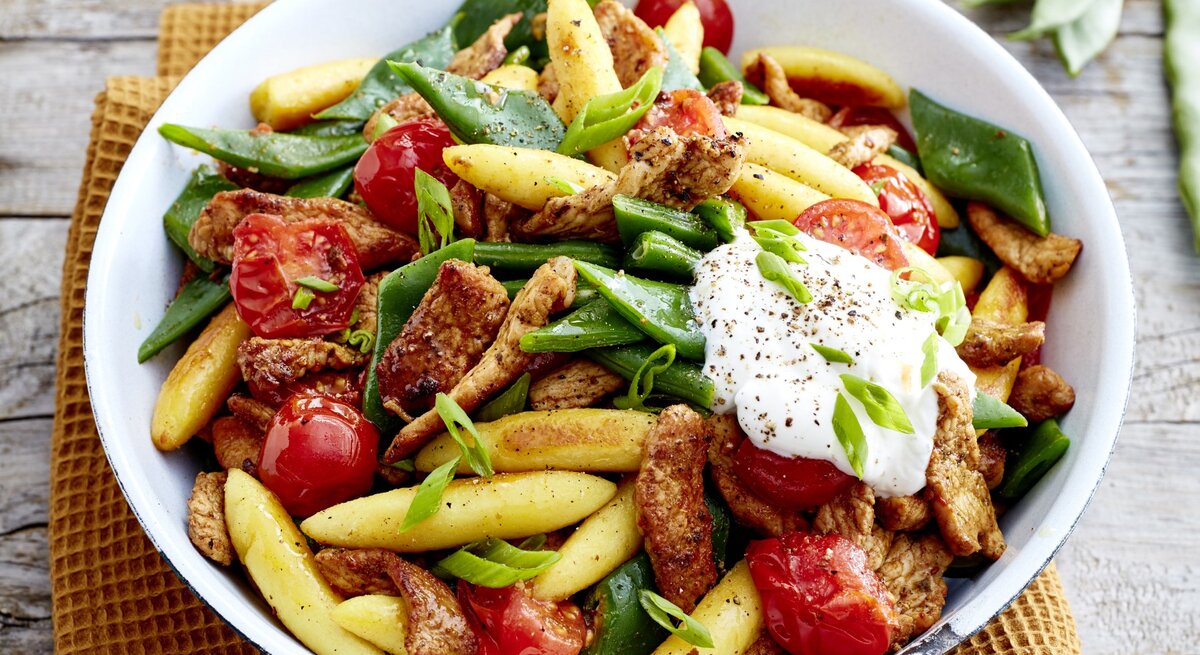 Gyros-Schupfnudel-Pfanne in weißem Teller auf einem gelben Küchentuch, Holzuntergrund und Bohnen im Hintergrund. 