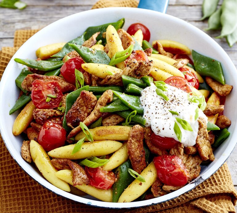 Gyros-Schupfnudel-Pfanne in weißem Teller auf einem gelben Küchentuch, Holzuntergrund und Bohnen im Hintergrund.  | © EAT CLUB / Isabell Triemer 