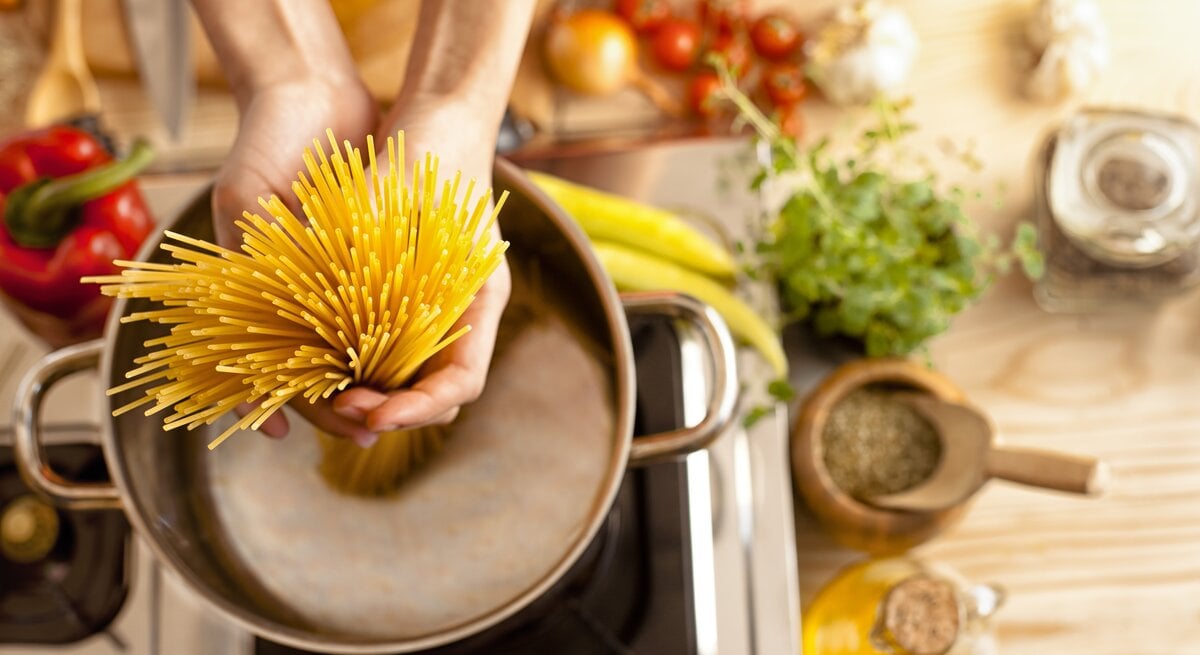Spaghetti werden in einen Topf voll Wasser gegeben. Um den Herd herum liegen verschiedene, typisch italienische Zutaten. 