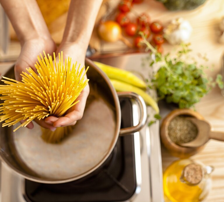 Spaghetti werden in einen Topf voll Wasser gegeben. Um den Herd herum liegen verschiedene, typisch italienische Zutaten.  | © iStock.com/zeljkosantrac