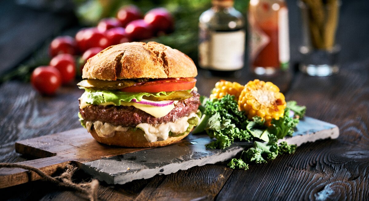 Ein veganer Elote-Burger mit Gemüse, weißer Maissoße, daneben liegen geschnittene Maiskolben und Salatblätter, im Hintergrund sieht man unscharf weitere Zutaten.
