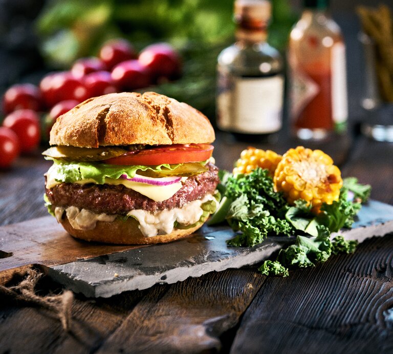 Ein veganer Elote-Burger mit Gemüse, weißer Maissoße, daneben liegen geschnittene Maiskolben und Salatblätter, im Hintergrund sieht man unscharf weitere Zutaten. | © Getty Images/ Olivia