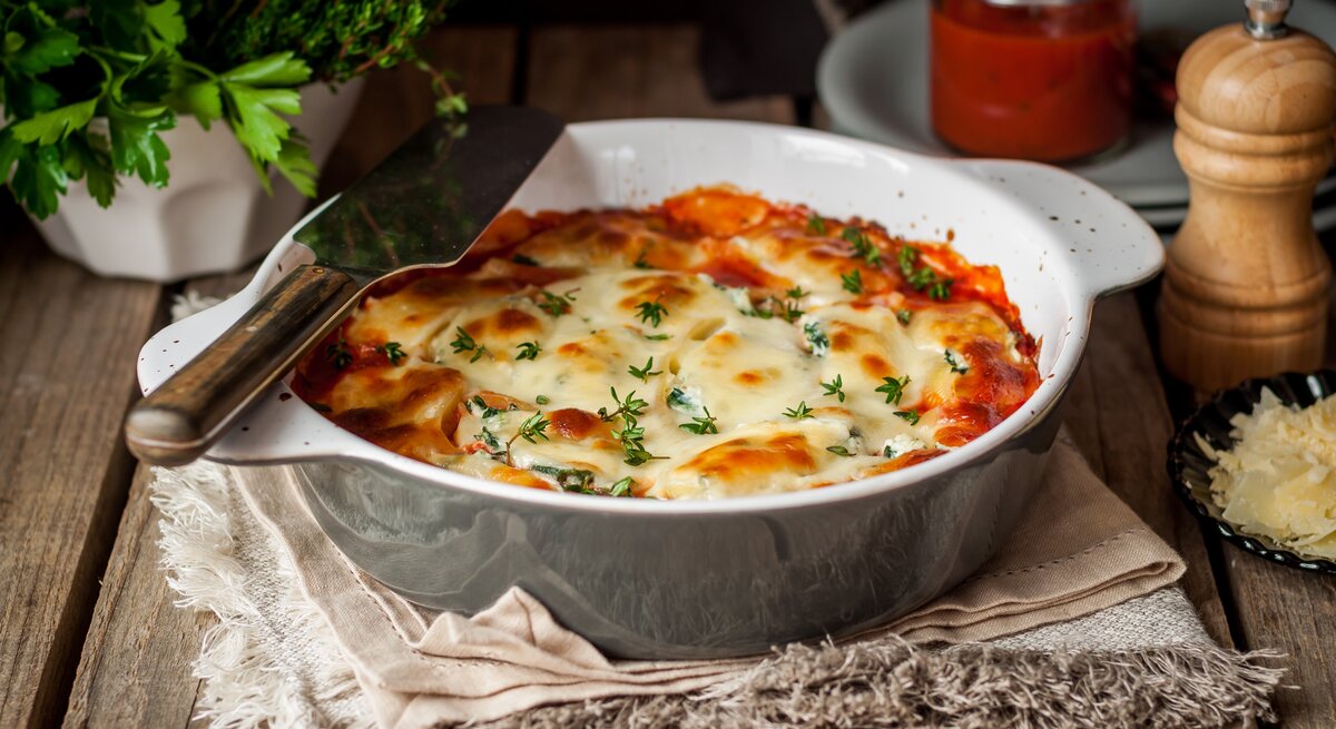 Garnelenauflauf mit Tagliatelle und 3-Käse-Béchamel in Auflaufform auf Tuch auf Holztisch. Daneben Kräuter, geriebener Käse, ein Glas mit Soße und Pfeffermühle. Frontalansicht.
