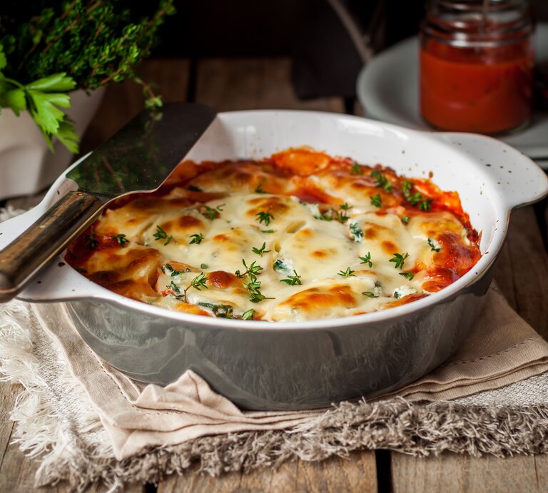 Garnelenauflauf mit Tagliatelle und 3-Käse-Béchamel in Auflaufform auf Tuch auf Holztisch. Daneben Kräuter, geriebener Käse, ein Glas mit Soße und Pfeffermühle. Frontalansicht. | © shutterstock / Tatiana Vorona