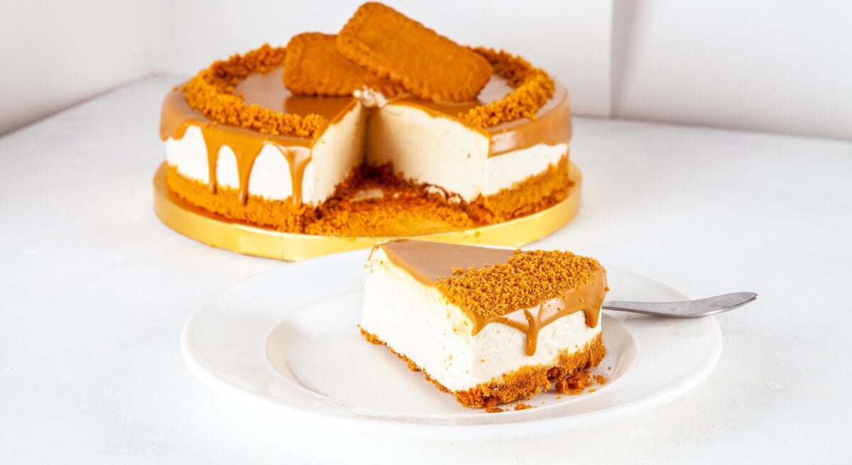 Goldene Tortenplatte, darauf ein Käsekuchen mit Lotus-Keksen. Ein Stück wurde herausgeschnitten und auf einem Teller angerichtet. 