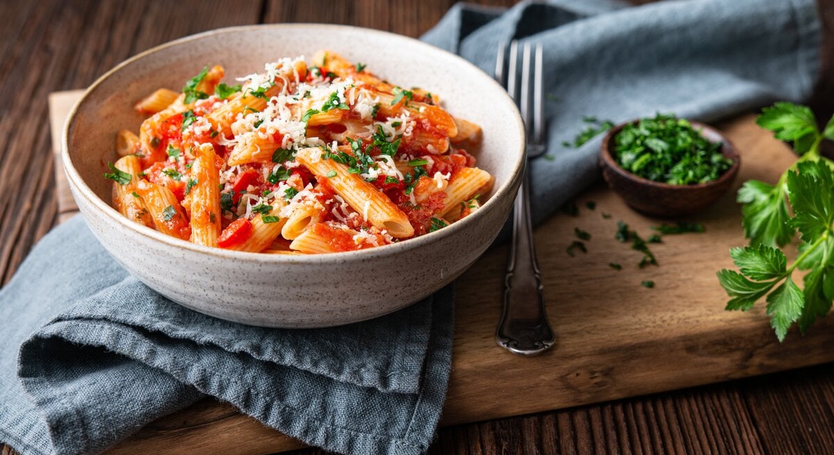 Frontalsicht: Eine Keramikschüssel steht auf einem Küchenhandtuch und dieses auf einem Holzbrett. In der Schüssel sind Nudeln mit einer Tomatensoße, Petersilie und Parmesan. das Gericht heißt Penne Cinque Pi. Neben der Schüssel liegt eine Gabel. Neben der ist eine kleine Schüssel mit frischer Petersilie.