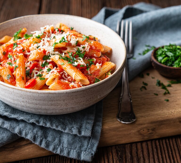 Frontalsicht: Eine Keramikschüssel steht auf einem Küchenhandtuch und dieses auf einem Holzbrett. In der Schüssel sind Nudeln mit einer Tomatensoße, Petersilie und Parmesan. das Gericht heißt Penne Cinque Pi. Neben der Schüssel liegt eine Gabel. Neben der ist eine kleine Schüssel mit frischer Petersilie. | © Shutterstock /  Marcus Z-pics