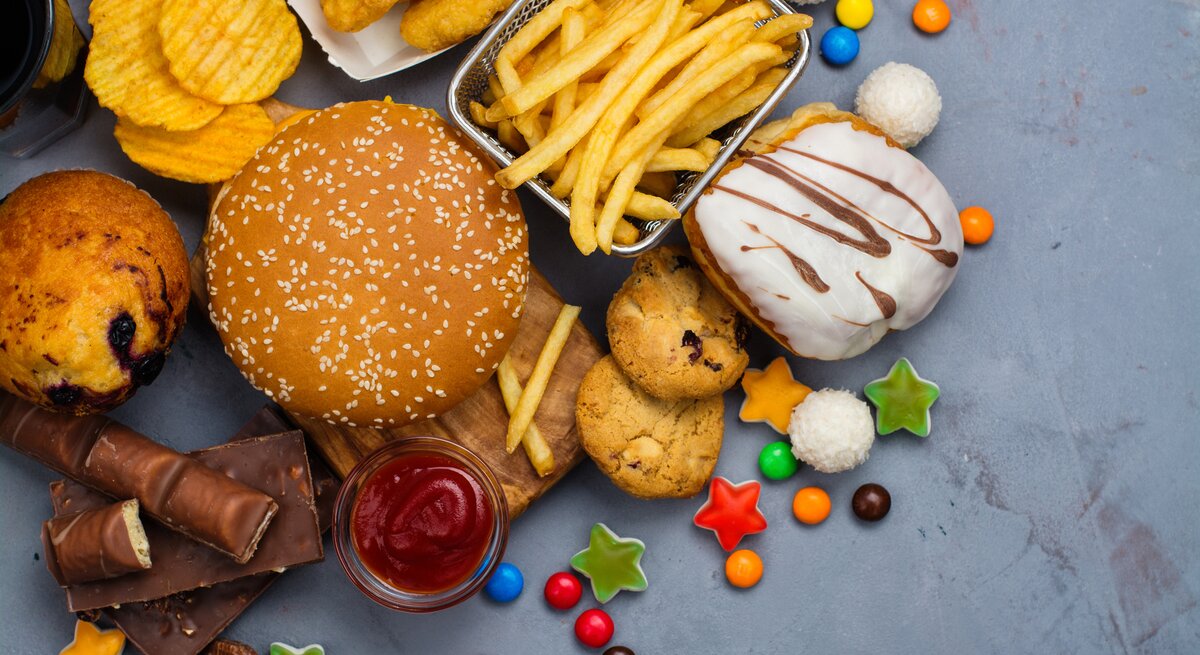 Pommes, Burger, Gummitierchen und Kuchen liegen auf einem Tisch. Diese Lebensmittel fördern eine Fettleber. 