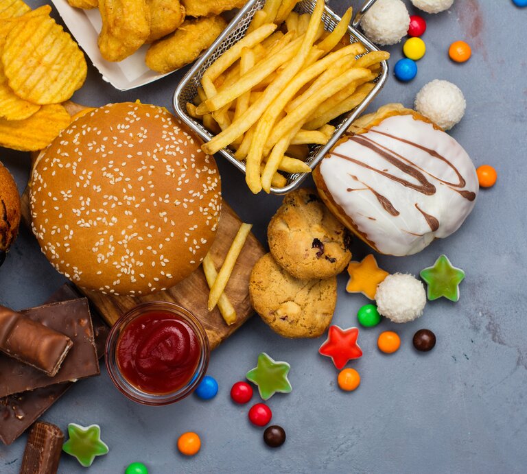 Pommes, Burger, Gummitierchen und Kuchen liegen auf einem Tisch. Diese Lebensmittel fördern eine Fettleber.  | © GettyImages/happy_lark