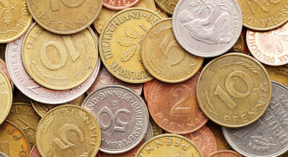 Ein Haufen verschiedener Pfennig-Münzen, die in Größe, Farbe und Design variieren. Einige zeigen Zahlen wie "10" oder "5", was auf ihren Nennwert in Pfennig hinweist. Einige Münzen zeigen Blätter und Ähren, während eine silberne Münze in der Mitte das Relief eines Mannes zeigt, der mit einem Hammer auf einem Amboss schlägt. Die Münzen scheinen aus verschiedenen Metallen zu bestehen, einige sind goldfarben, andere kupferfarben und wieder andere silberfarben. Sie sind zufällig angeordnet und überlappen sich teilweise.