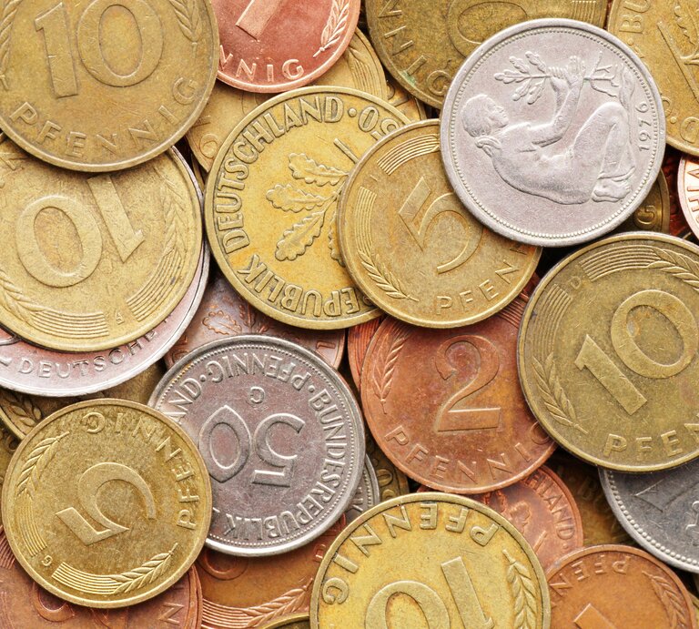 Ein Haufen verschiedener Pfennig-Münzen, die in Größe, Farbe und Design variieren. Einige zeigen Zahlen wie "10" oder "5", was auf ihren Nennwert in Pfennig hinweist. Einige Münzen zeigen Blätter und Ähren, während eine silberne Münze in der Mitte das Relief eines Mannes zeigt, der mit einem Hammer auf einem Amboss schlägt. Die Münzen scheinen aus verschiedenen Metallen zu bestehen, einige sind goldfarben, andere kupferfarben und wieder andere silberfarben. Sie sind zufällig angeordnet und überlappen sich teilweise. | © imago images / Panthermedia