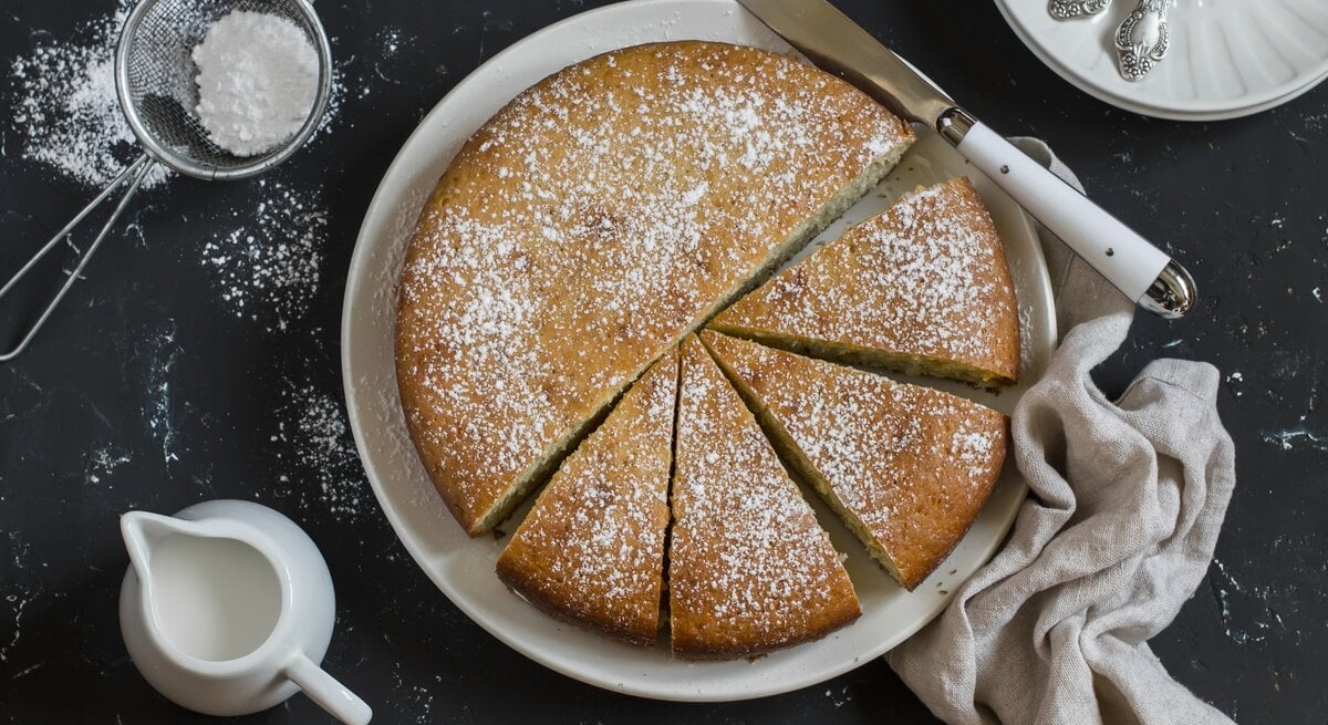 Runder, heller Kuchen bestäubt mit Puderzucker auf weißem Teller. Dieser steht auf einer dunklen Arbeitsfläche. 