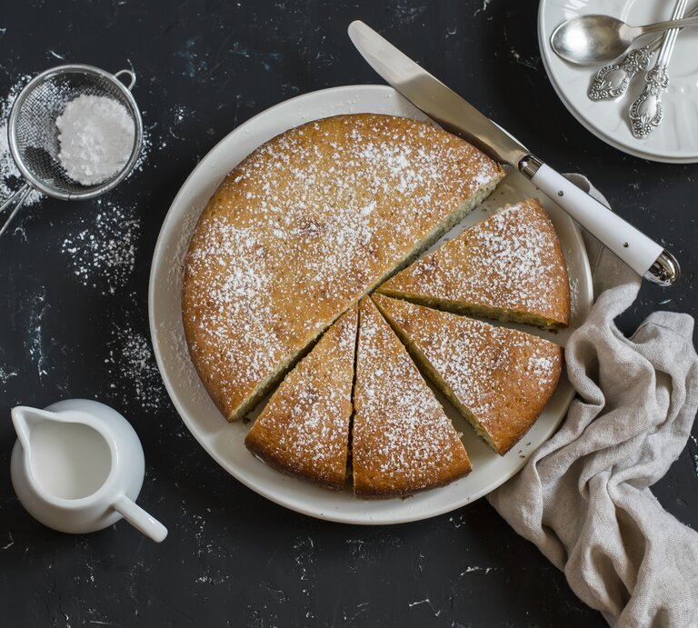 Runder, heller Kuchen bestäubt mit Puderzucker auf weißem Teller. Dieser steht auf einer dunklen Arbeitsfläche.  | © Getty Images/OksanaKiian