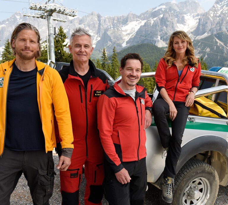 Vier "Die Bergretter"-Darsteller stehen in Rettungsdienstuniformen neben einem Offroad-Rettungsfahrzeug mit der Aufschrift "Bergrettung". Im Hintergrund sind Berggipfel zu sehen. Von links nach rechts: Sebastian Ströbel als Markus Kofler, Robert Lohr als Michael "Michi" Dörfler, Markus Brandl als Tobias Herbrechter und Luise Bähr als Katharina Strasser. | © ZDF/Barbara Bauriedl
