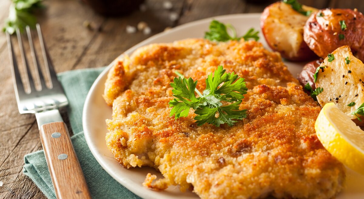 Ein Schnitzel auf einem Teller mit Ofenkartoffeln, Petersilie und Zitrone. Alles auf einem rustikalen Holztisch mit dunkelgrüner Tischdecke und Gabel. Sehr nah von der Seite fotografiert.