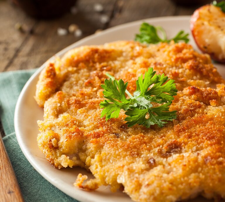 Ein Schnitzel auf einem Teller mit Ofenkartoffeln, Petersilie und Zitrone. Alles auf einem rustikalen Holztisch mit dunkelgrüner Tischdecke und Gabel. Sehr nah von der Seite fotografiert. | © Adobe Stock/ Brent Hofacker
