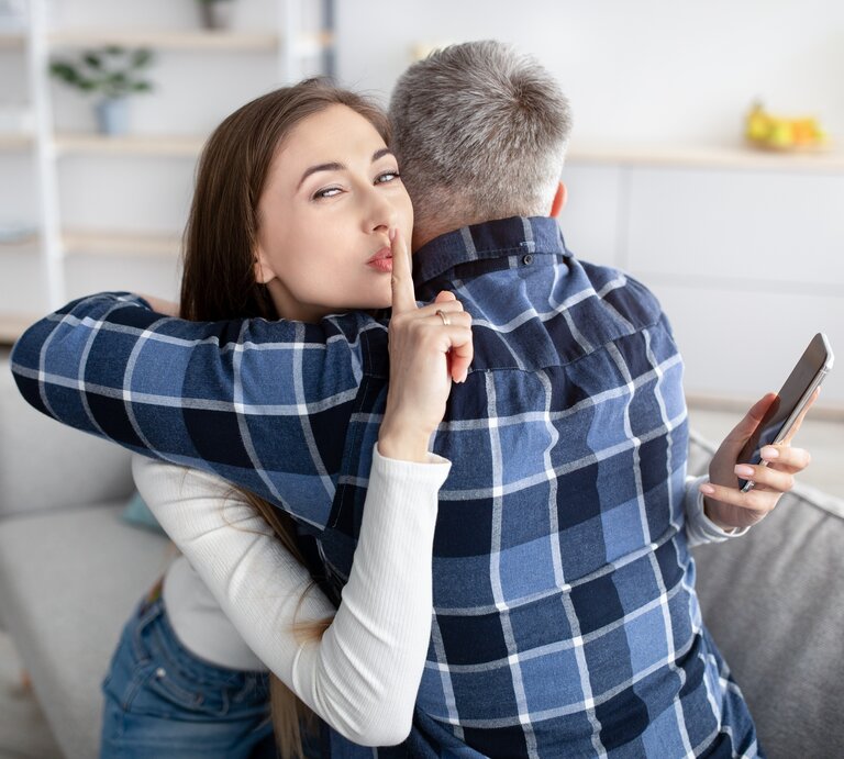 Frau und Mann umarmen sich und die Frau hat hinter dem Rücken des Mannes ein Handy in der Hand, guckt in die Kamera und macht das Hush-Zeichen. | © Shutterstock/Prostock-studio