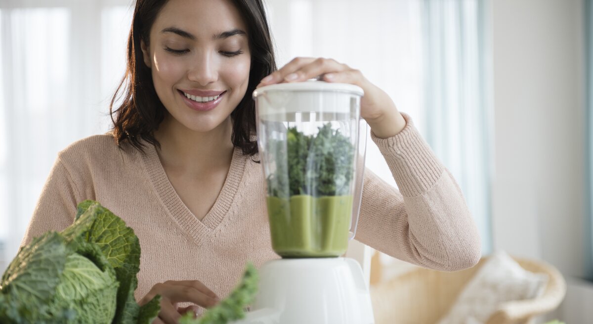 Eine lächelnde junge Frau bereitet in ihrer Küche einen grünen Smoothie zu. Sie legt ihre Hand auf den Deckel eines Mixers, der mit grünen Blättern gefüllt ist, vermutlich Kale oder Spinat. Vor ihr auf dem Tisch liegen frische grüne Gemüsesorten. Das Bild vermittelt ein Gefühl von Gesundheit und Wohlbefinden durch die Zubereitung von frischen, nahrhaften Lebensmitteln. Die Küche ist hell und einladend mit einem Fenster im Hintergrund.