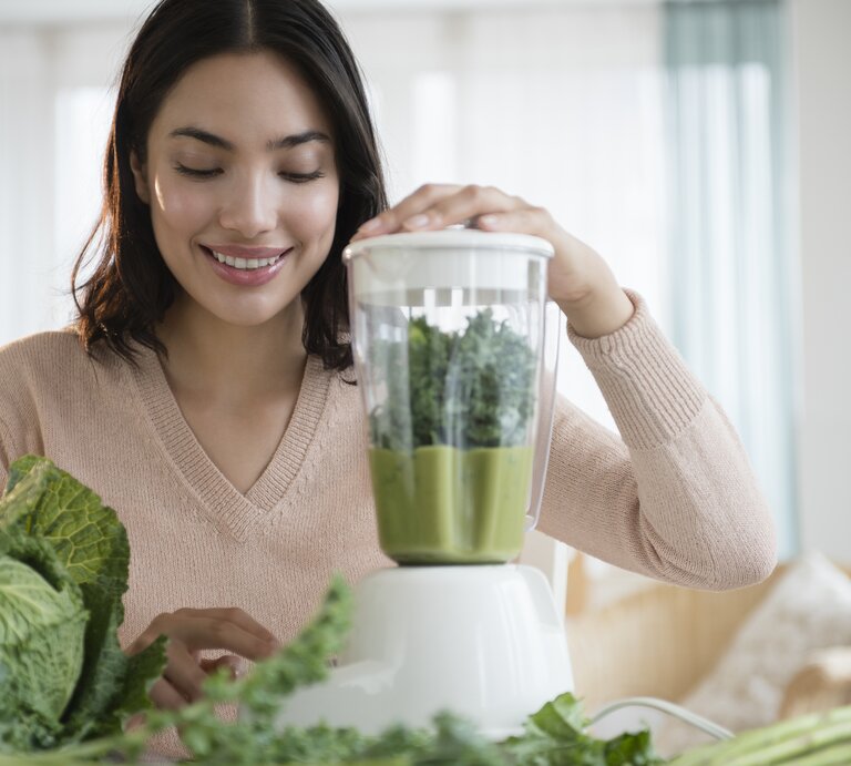 Eine lächelnde junge Frau bereitet in ihrer Küche einen grünen Smoothie zu. Sie legt ihre Hand auf den Deckel eines Mixers, der mit grünen Blättern gefüllt ist, vermutlich Kale oder Spinat. Vor ihr auf dem Tisch liegen frische grüne Gemüsesorten. Das Bild vermittelt ein Gefühl von Gesundheit und Wohlbefinden durch die Zubereitung von frischen, nahrhaften Lebensmitteln. Die Küche ist hell und einladend mit einem Fenster im Hintergrund. | © Getty Images/JGI/Jamie Grill