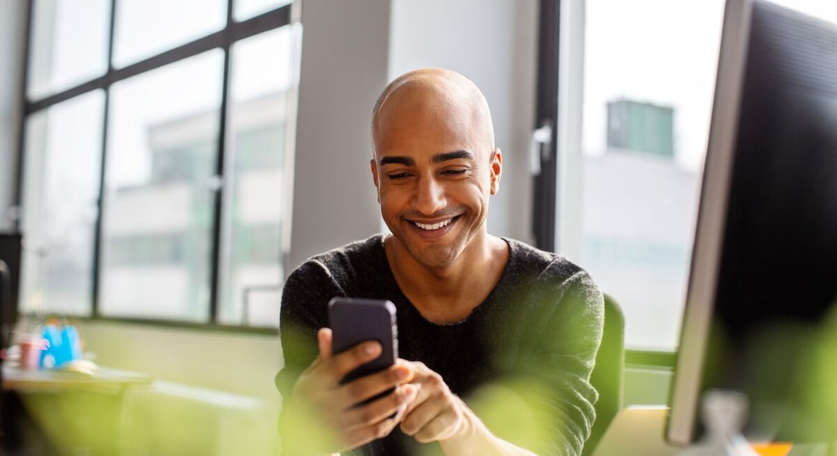 Junger Mann mit Glatze lacht am Schreibtisch beim Blick in sein Handy