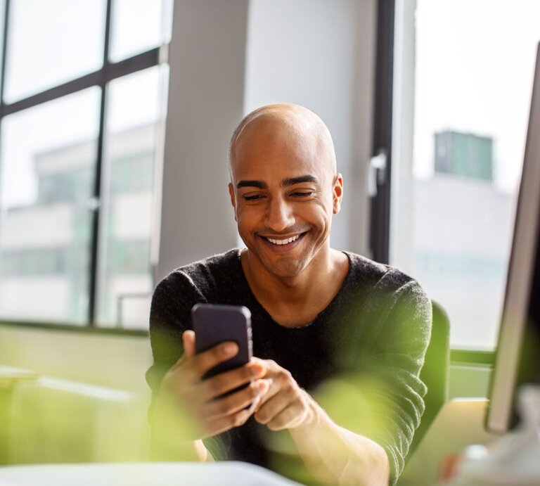 Junger Mann mit Glatze lacht am Schreibtisch beim Blick in sein Handy | © Getty Images / Luis Alvarez