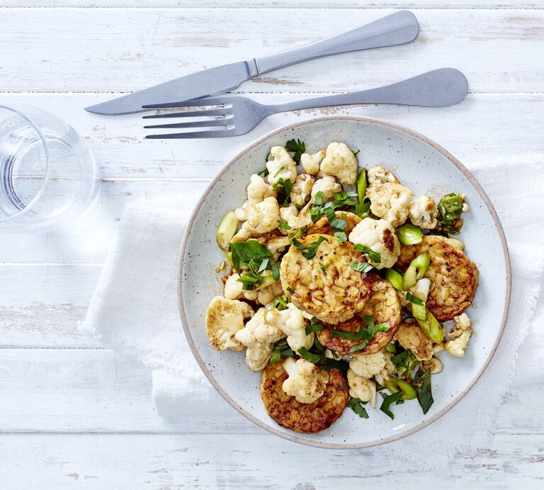 Gemüsepfanne mit Tempeh in einem hellblauen Teller auf einem Geschirrtuch neben einem Glas Wasser und Besteck auf einem weißen Tisch. | © EAT CLUB/Isabell Triemer