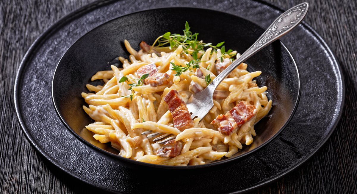 Schwarzer Teller mit Pasta mit Pancetta in Pecorino-Creme auf hölzernem Untergrund. 