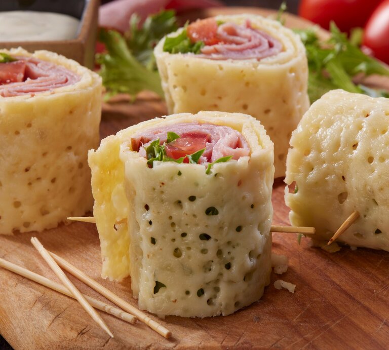 Mozzarella-Röllchen auf Holzbrett mit Zahnstochern. Im Hintergrund Salat, Tomaten und Dipschale. Frontalansicht. | © Getty Images / LauriPatterson