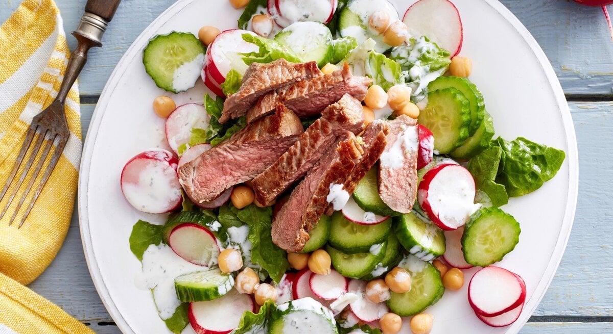 Auf einem Teller ist ein Zaziki-Salat mit Steakstreifen angerichtet. Er ist mit Radieschen und Gurke verfeinert.