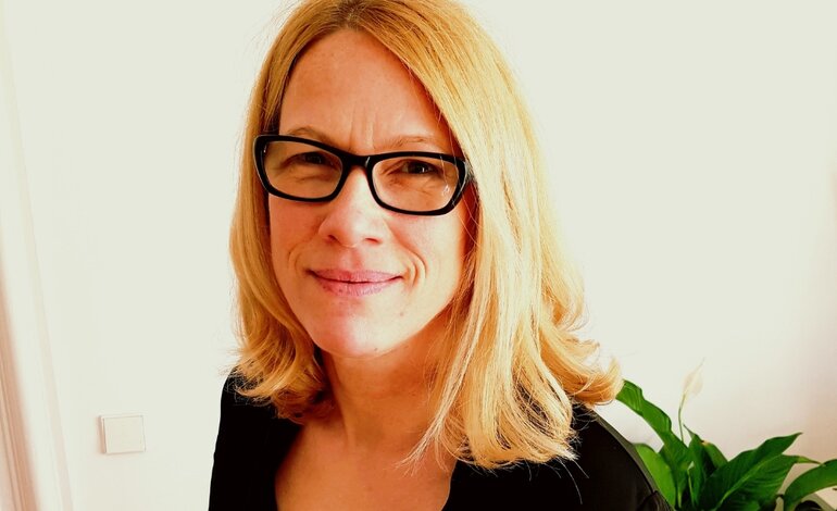 Das Bild zeigt eine lächelnde Frau mit blondem, schulterlangem Haar, die eine schwarze Brille trägt. Sie blickt direkt in die Kamera und hat einen freundlichen Gesichtsausdruck. Sie ist in einem Innenraum vor einem Hintergrund, der teilweise eine weiße Wand und eine grüne Pflanze zeigt, was eine häusliche oder büroähnliche Umgebung suggeriert. Die Frau trägt eine schwarze Kleidung, was auf einen formellen oder beruflichen Kontext hindeuten könnte. | © privat