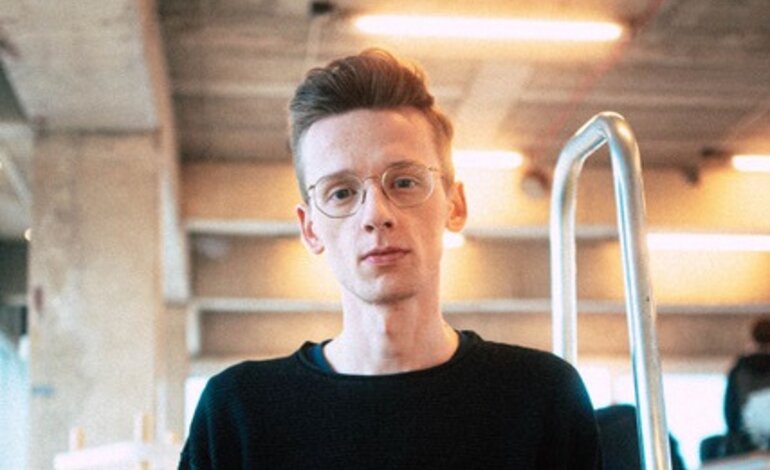 Ein junger Mann mit Brille und ordentlichem Haarschnitt trägt einen schwarzen Pullover und steht in einem Innenraum, der wie ein modernes Büro oder Café wirkt, mit unscharfem Hintergrund, der Bewegung und Geschäftigkeit suggeriert. | © privat