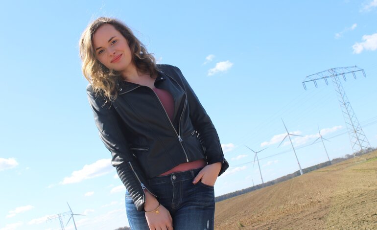 Das Bild zeigt eine junge Frau, die draußen steht und selbstbewusst in die Kamera lächelt. Sie trägt eine schwarze Lederjacke, ein dunkles Oberteil und Jeans. Im Hintergrund ist eine ländliche Landschaft mit einem klaren blauen Himmel, Windrädern und einem Strommast zu sehen. Die Pose der Frau ist entspannt und lässig, und sie scheint die frische Luft und die Umgebung zu genießen. | © privat