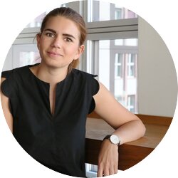 Eine junge Frau mit zurückgebundenen Haaren, einem schwarzen Oberteil und einer Uhr sitzt lächelnd vor einem Fenster, durch das man auf urbane Architektur blicken kann, was ein entspanntes und professionelles Ambiente vermittelt. | © Mandy Buchholz