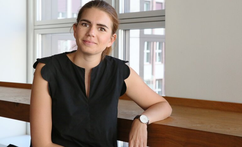 Eine junge Frau mit zurückgebundenen Haaren, einem schwarzen Oberteil und einer Uhr sitzt lächelnd vor einem Fenster, durch das man auf urbane Architektur blicken kann, was ein entspanntes und professionelles Ambiente vermittelt. | © Mandy Buchholz