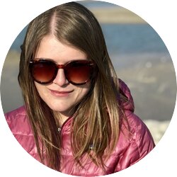 Das Bild zeigt eine Frau mit blonden Haaren und Sonnenbrille, die in die Kamera lächelt. Sie trägt eine pinke Jacke und scheint sich an einem Strand oder einer Küstenregion zu befinden, erkennbar an dem Sand und Wasser im Hintergrund. Die Sonne scheint auf ihr Gesicht, was ihr einen fröhlichen Ausdruck verleiht. | © privat