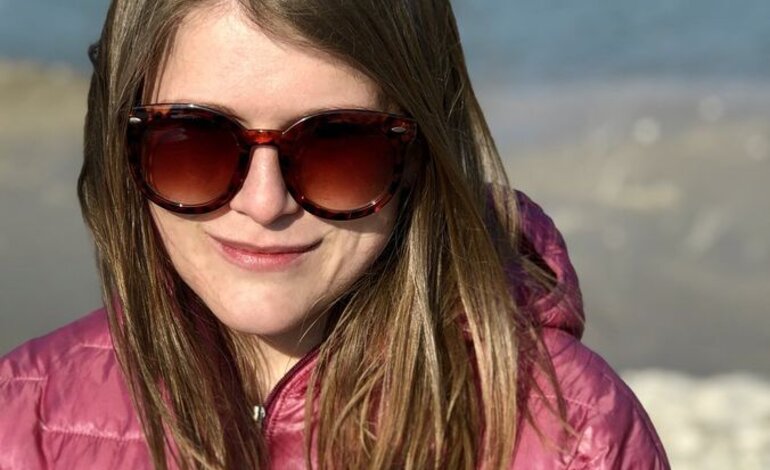 Das Bild zeigt eine Frau mit blonden Haaren und Sonnenbrille, die in die Kamera lächelt. Sie trägt eine pinke Jacke und scheint sich an einem Strand oder einer Küstenregion zu befinden, erkennbar an dem Sand und Wasser im Hintergrund. Die Sonne scheint auf ihr Gesicht, was ihr einen fröhlichen Ausdruck verleiht. | © privat