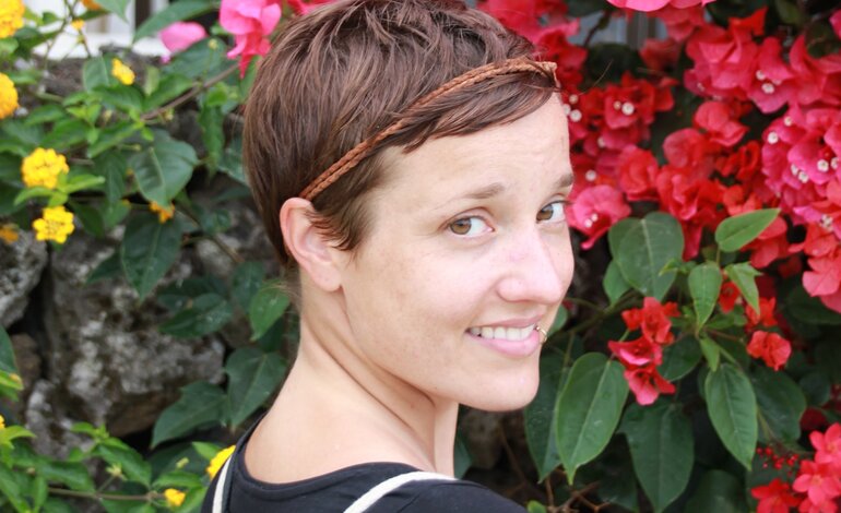 Eine Frau mit kurzen Haaren steht vor einer blumigen Kulisse und lächelt in die Kamera.  | © privat