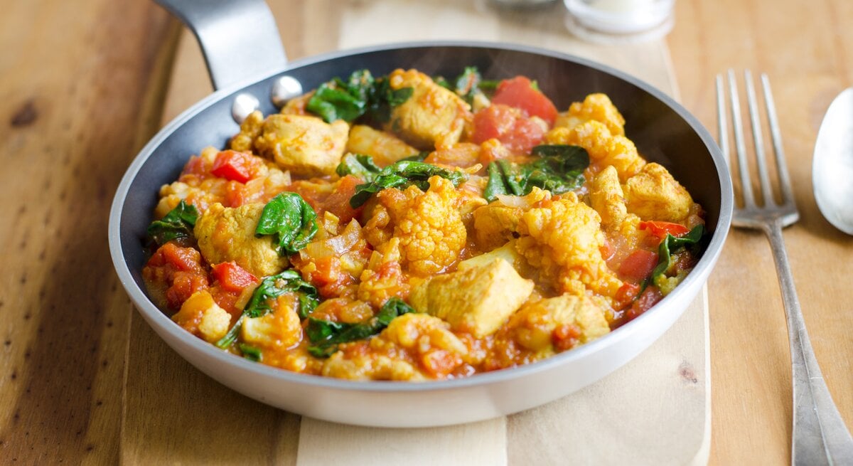 Schmackhaftes Hühnchen-Curry mit Tomaten und Spinat, serviert in einer Pfanne auf einem Holzbrett.