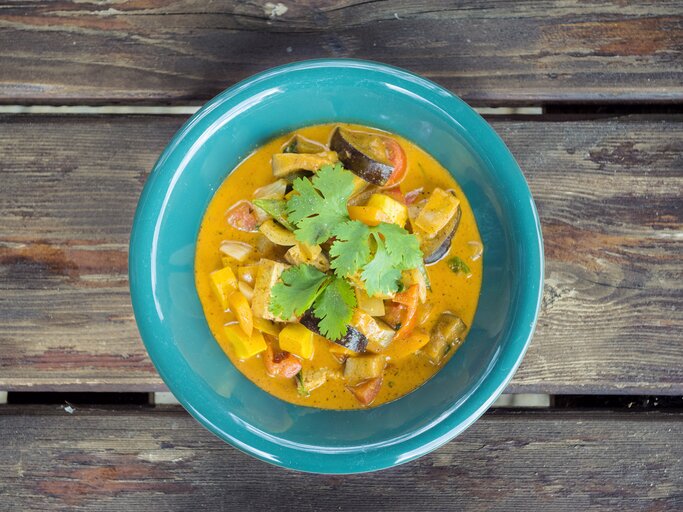 Ein grüner Teller mit würzigem Curry aus Gemüse und Koriander auf einem rustikalen Holztisch. | © iStock / Rosa Lazic