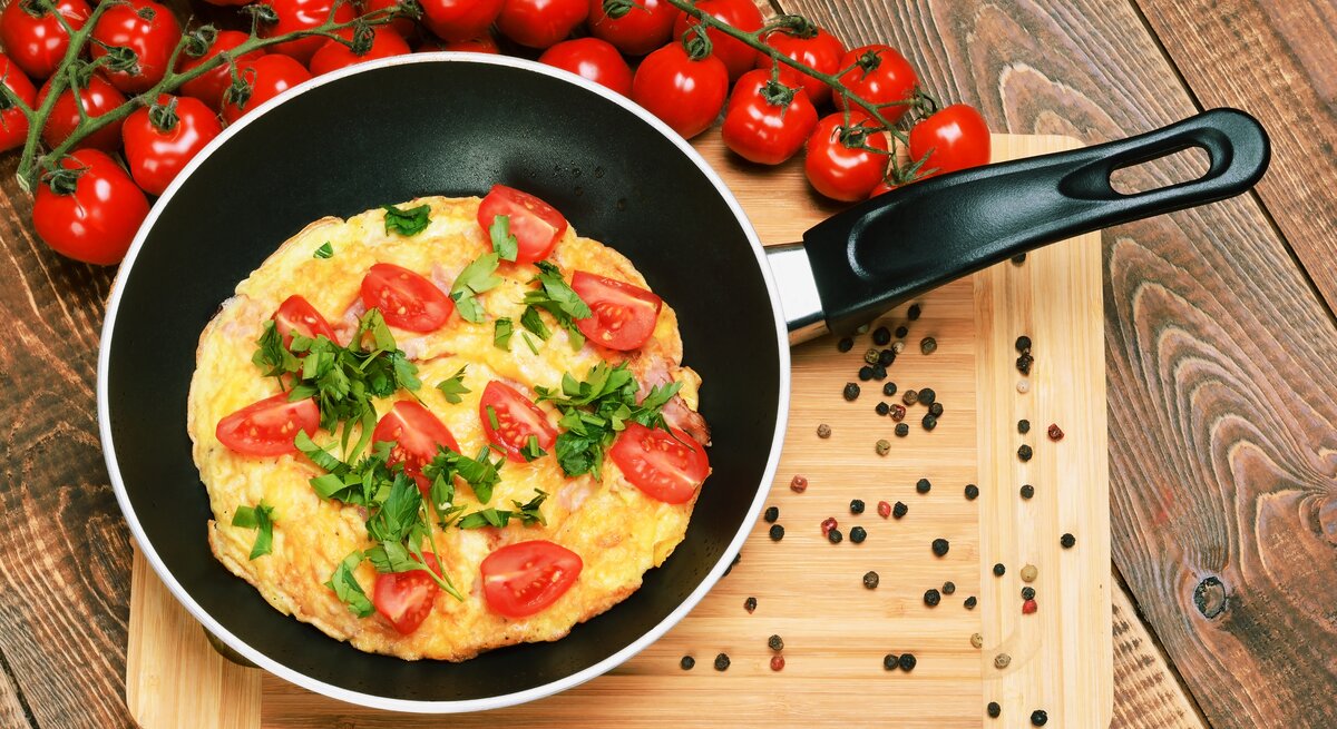 Eine Pfanne mit dem Tomatenomelett auf einem Holztisch mit einem Strauch Kirschtomaten von oben fotografiert.