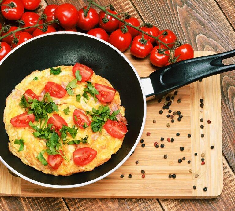 Eine Pfanne mit dem Tomatenomelett auf einem Holztisch mit einem Strauch Kirschtomaten von oben fotografiert. | © iStock.com/ madaland
