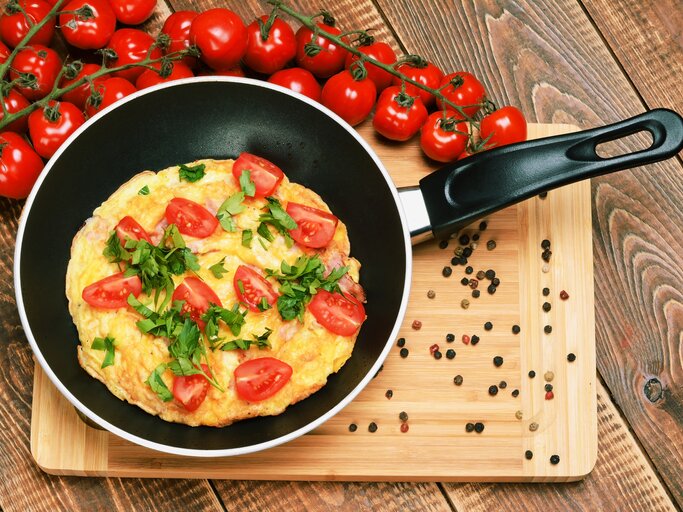 Eine Pfanne mit dem Tomatenomelett auf einem Holztisch mit einem Strauch Kirschtomaten von oben fotografiert. | © iStock.com/ madaland