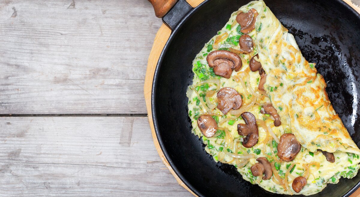 Eine Pfanne mit dem Pilz-Omelette auf einem hellen Holztisch direkt von oben fotografiert.